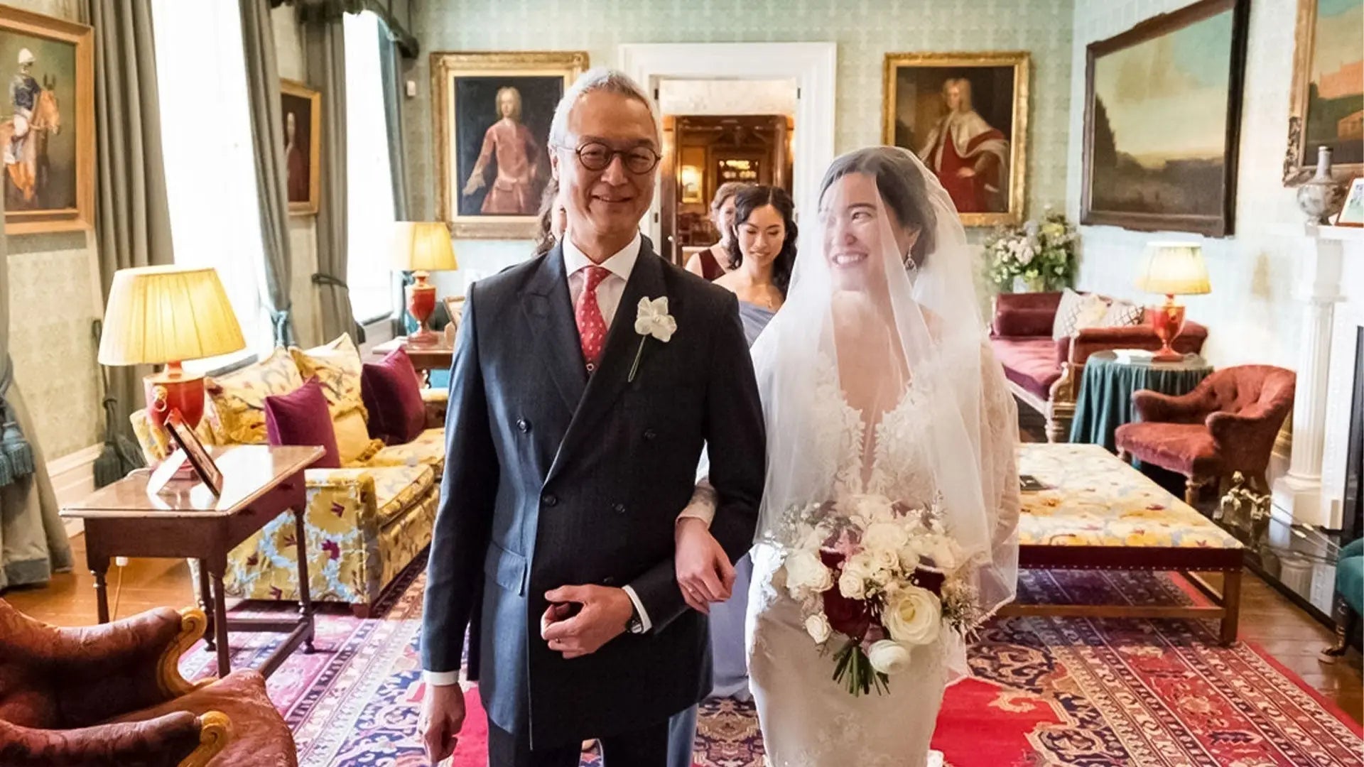 Father of the bride in a navy pinstripe suit and coral tie walking with the bride through a country house drawing room