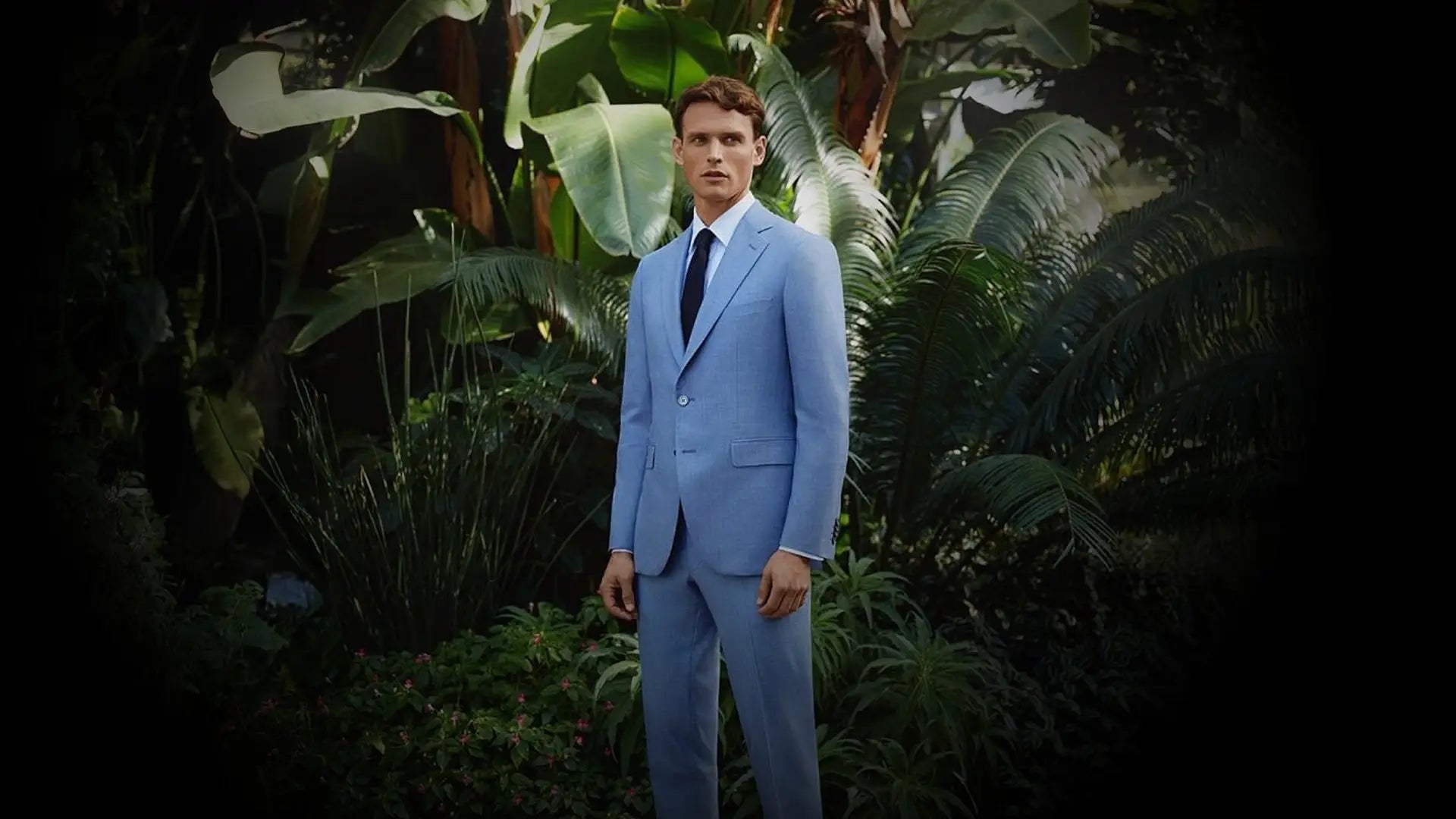 Man in a light blue suit with a white shirt and navy tie standing among tropical foliage