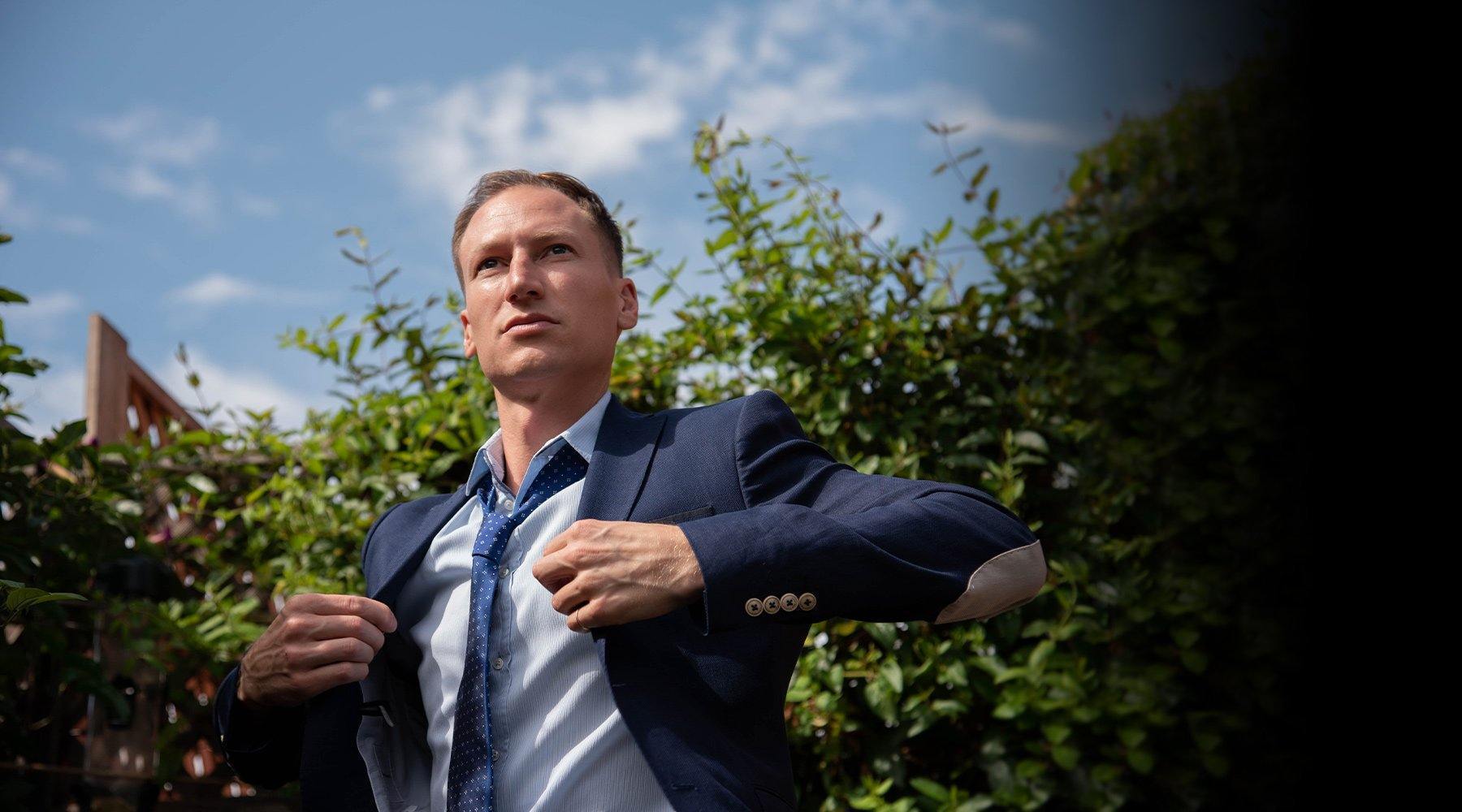 Man adjusting the lapels of a navy jacket outdoors against a hedge and open sky