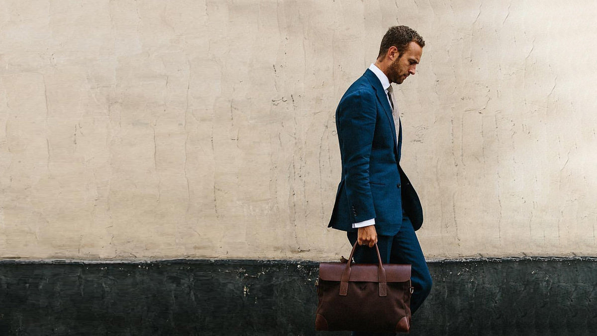 Man in a navy suit walking with a brown leather holdall past a plaster wall