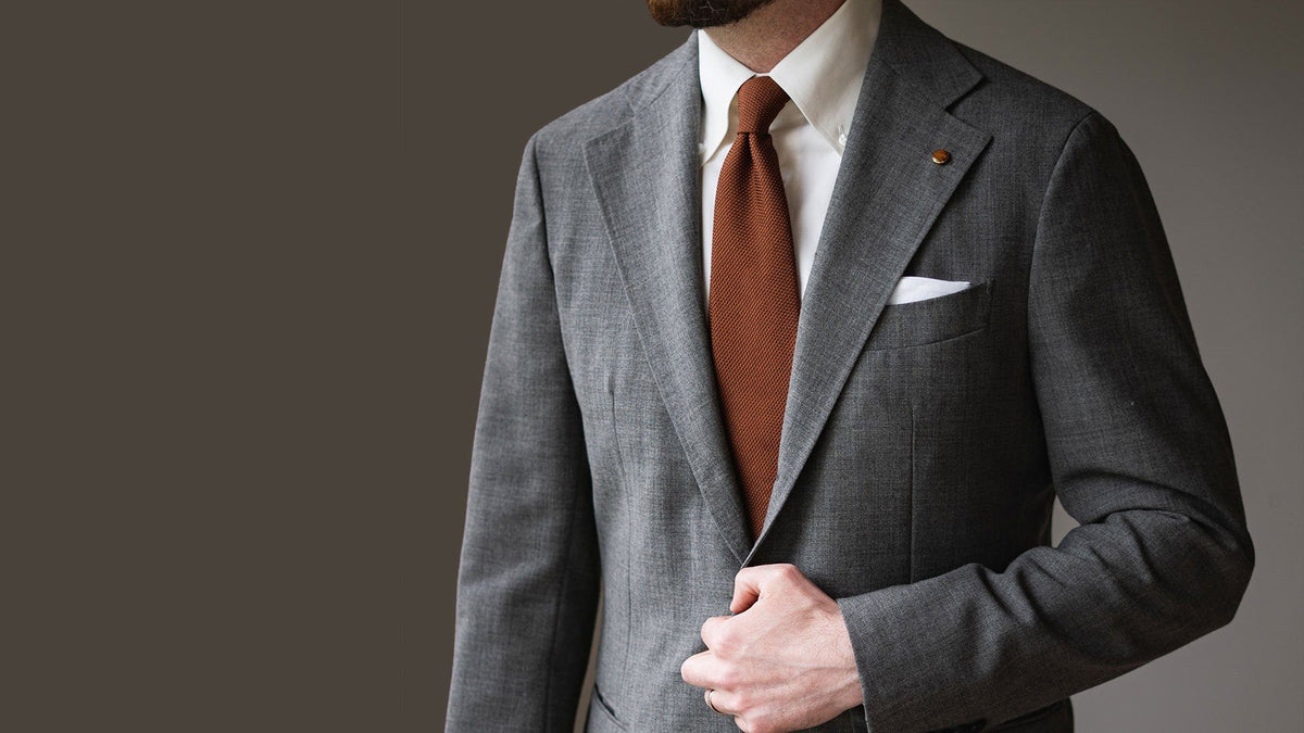 Grey glen check suit with a white shirt, burnt orange grenadine tie and white pocket square