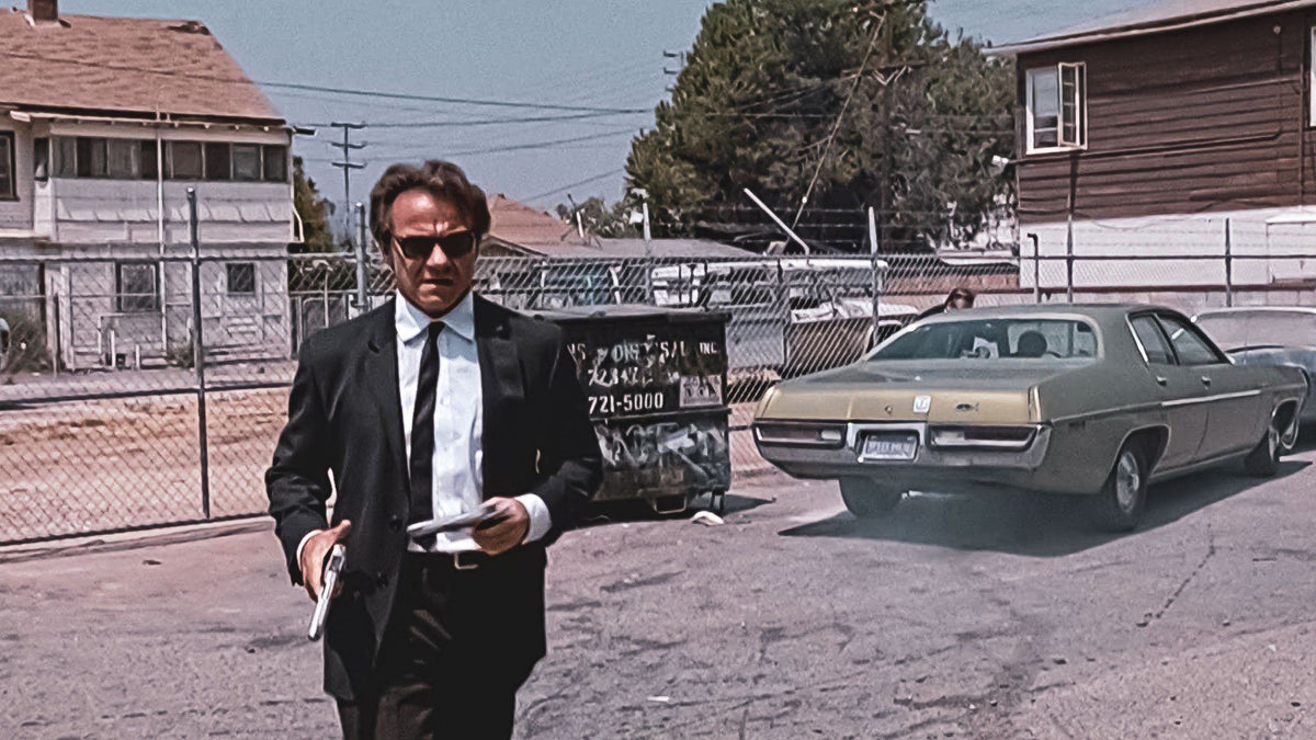 Man in a black suit, white shirt and black tie wearing sunglasses in a sunlit lot beside a vintage car