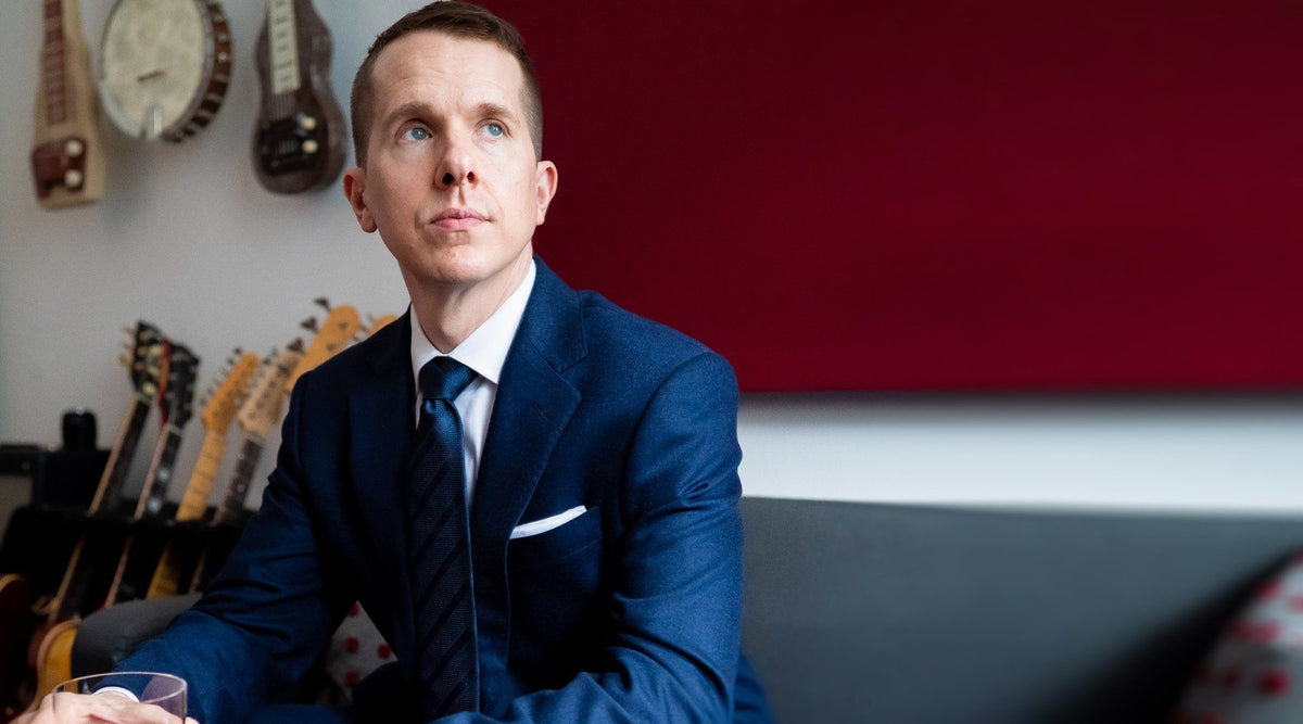 Man in a navy suit and striped navy tie seated in front of a wall of guitars