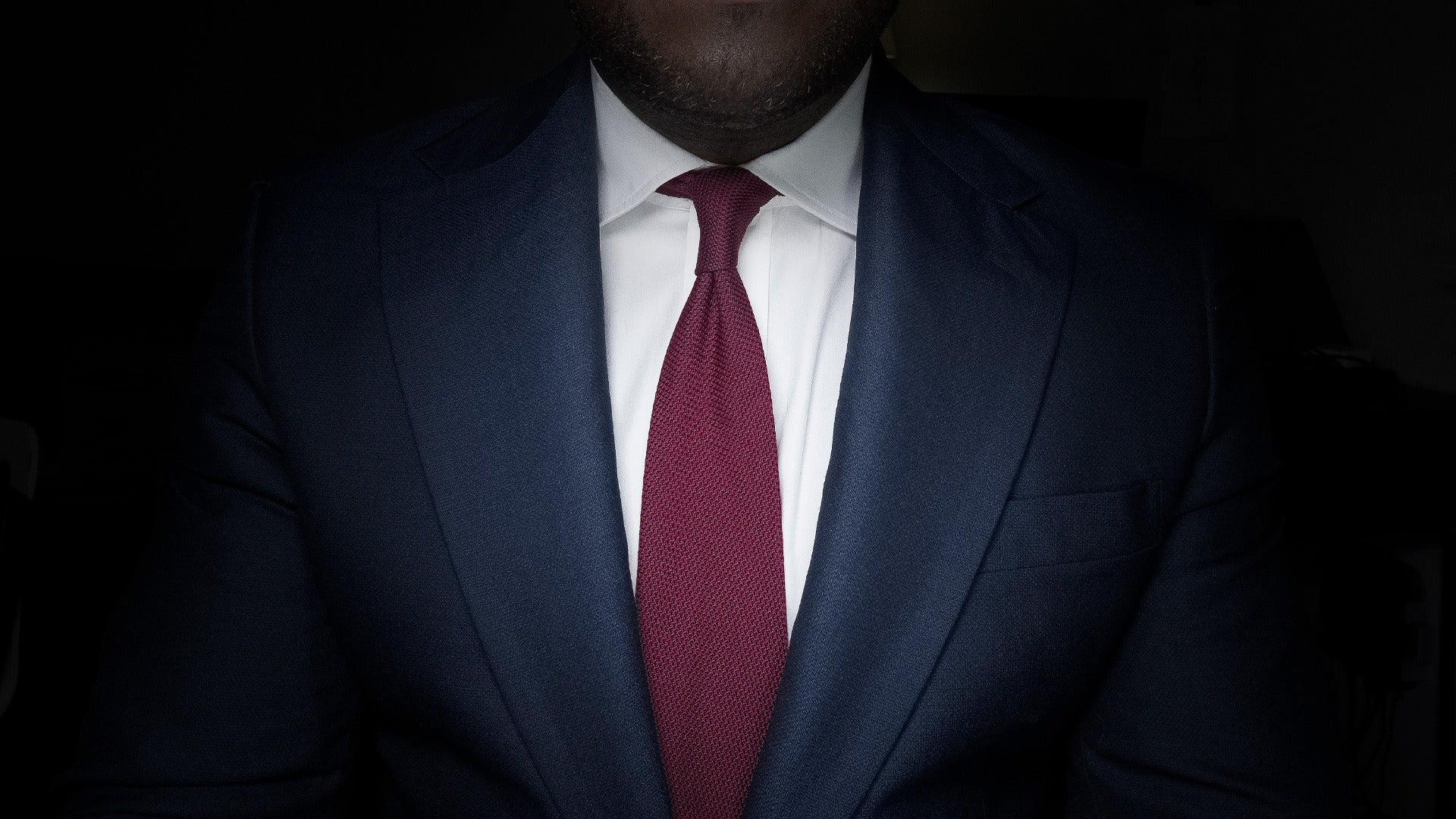 Burgundy grenadine tie in a four-in-hand knot, worn with a navy suit and white shirt, the open weave picked out by a single light.