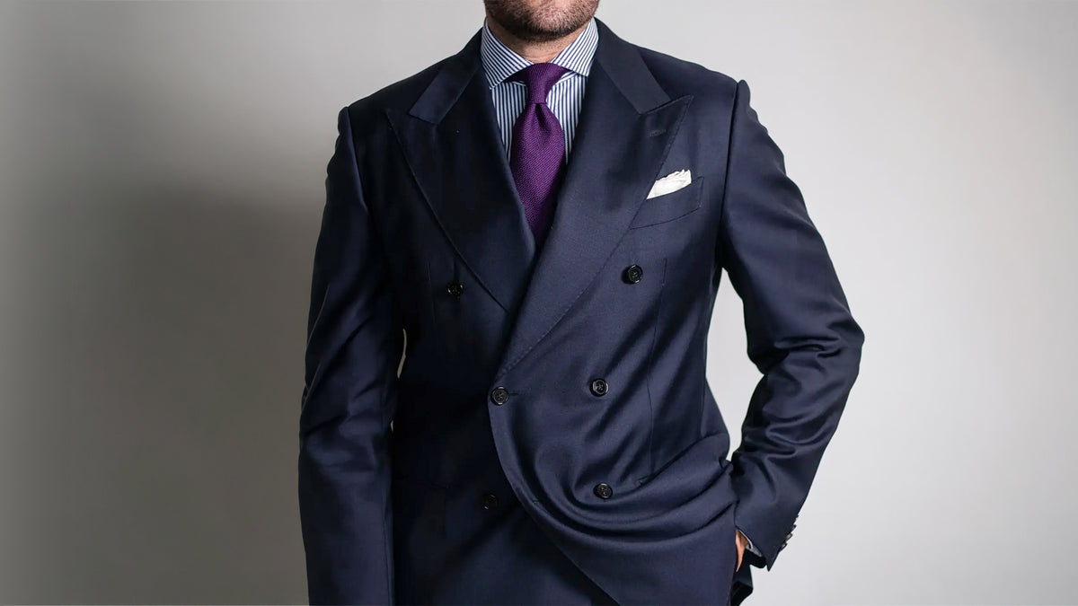 Purple grenadine tie in a four-in-hand knot, worn with a navy double-breasted suit, a blue and white striped shirt, and a white pocket square.