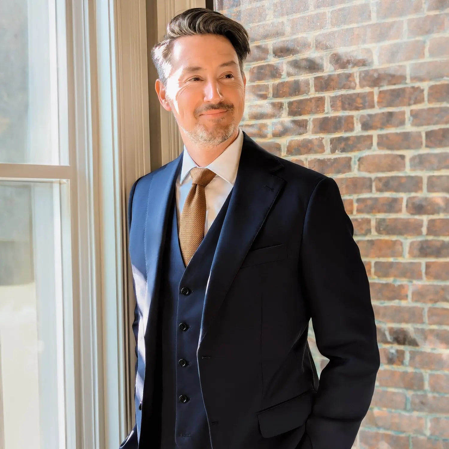 Man in a navy three-piece suit and tan tie standing by a window against a brick wall