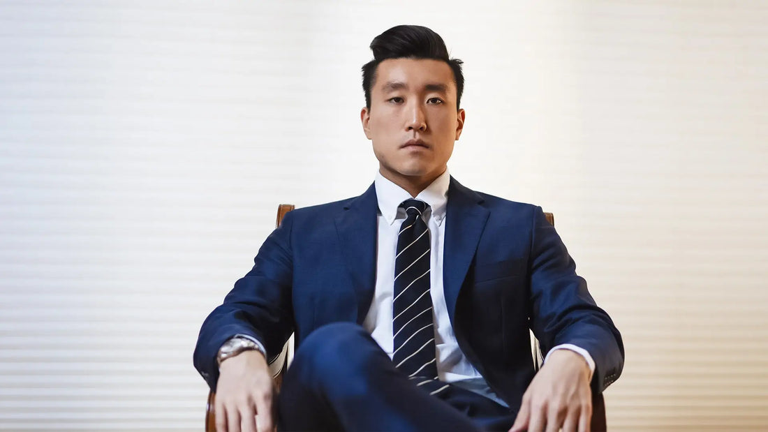 Man in a navy suit and navy striped tie seated against a pale background