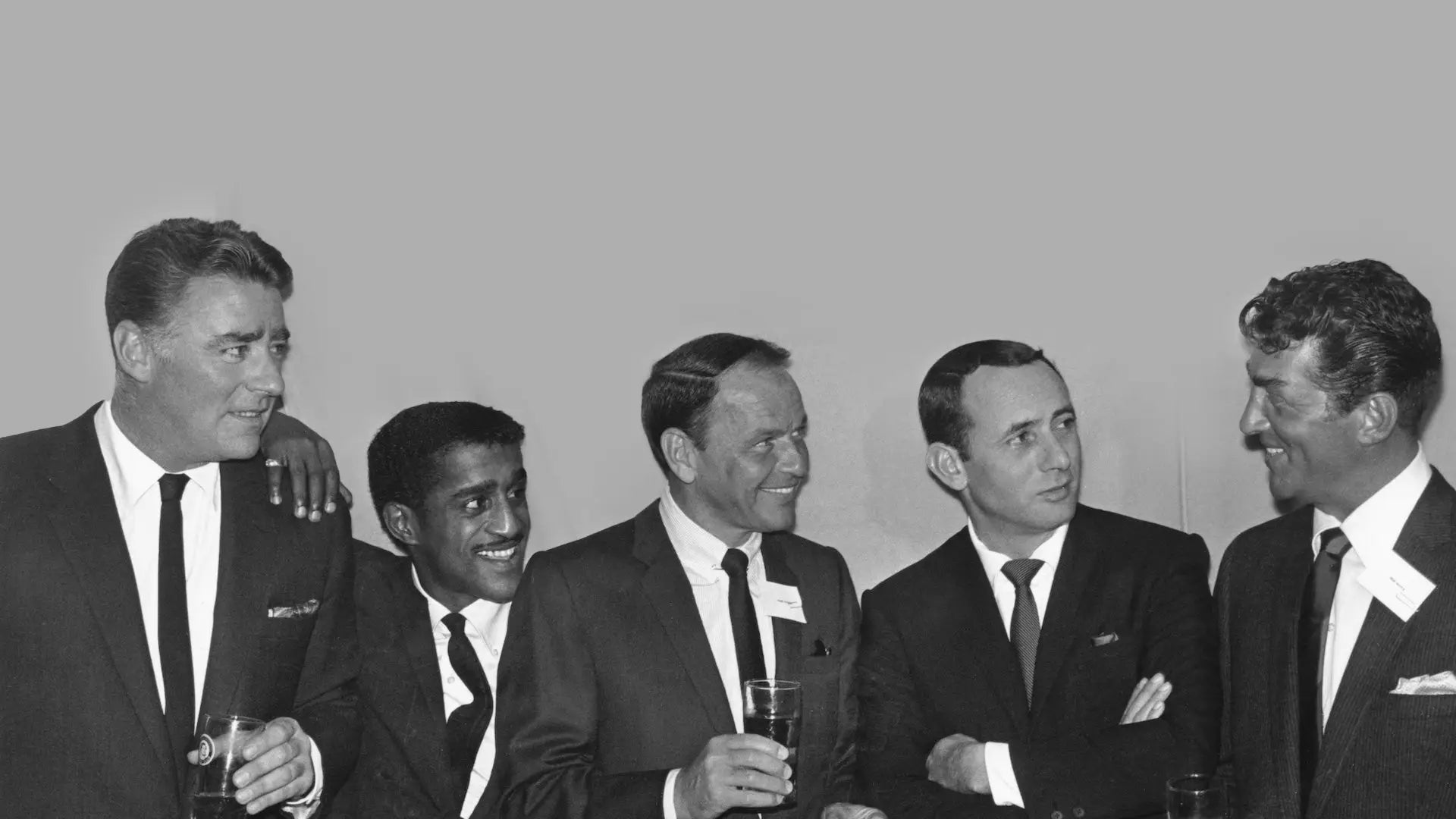 Black and white photograph of the Rat Pack in dark suits and black ties, holding drinks