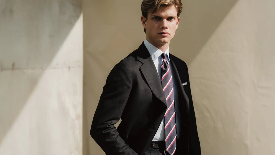 Young man in a black suit and navy and red striped tie against a sunlit wall