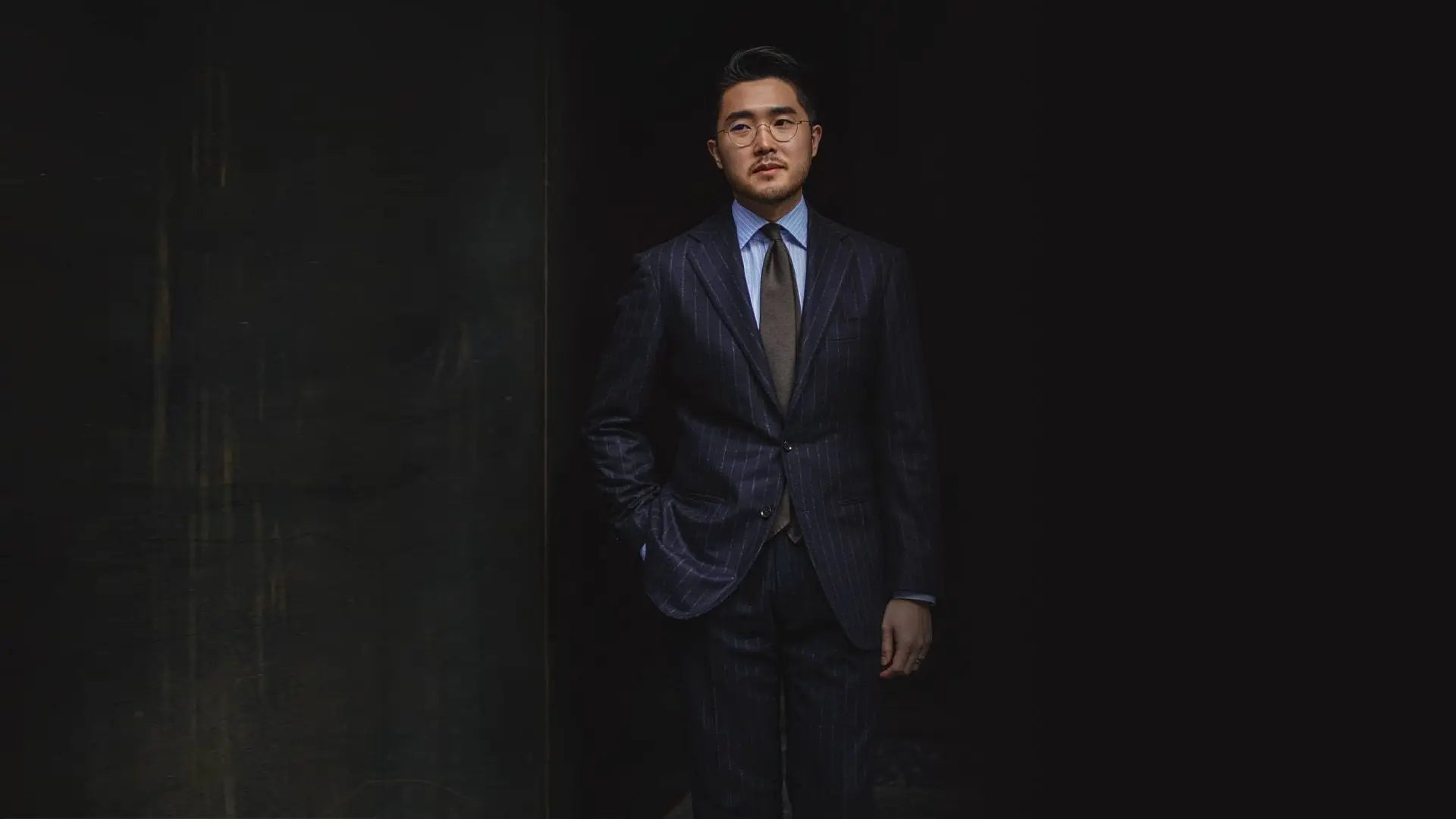 Man in a navy pinstripe suit with a blue shirt and brown tie against a dark wall