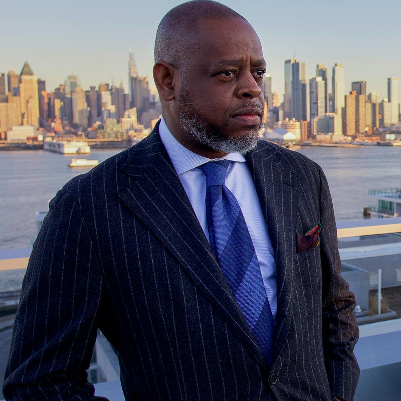 Close-up of man in pinstripe suit wearing navy and steel blue striped tie, city skyline in view.