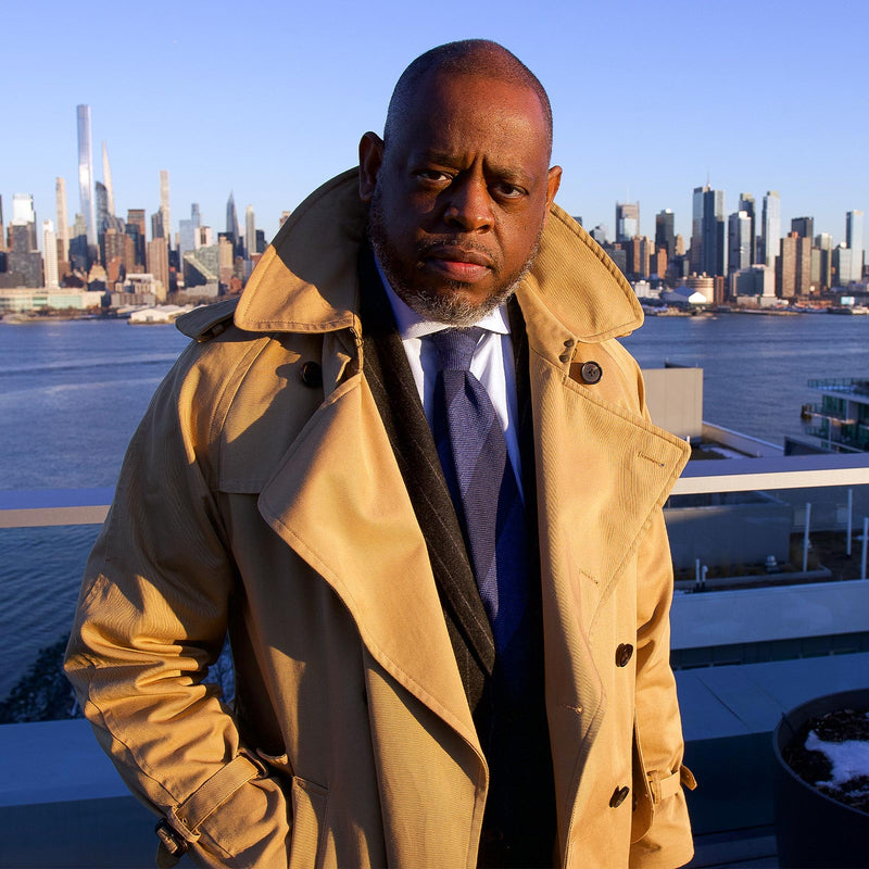 Man wearing a navy and steel blue striped tie with a trench coat, city skyline in the background.