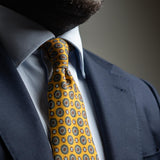 Close-up of medallion foulard pattern on golden yellow silk tie paired with light blue and navy blue suit.