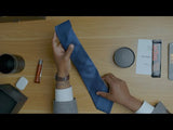 Hands holding a navy and steel blue block striped tie on a wooden table.